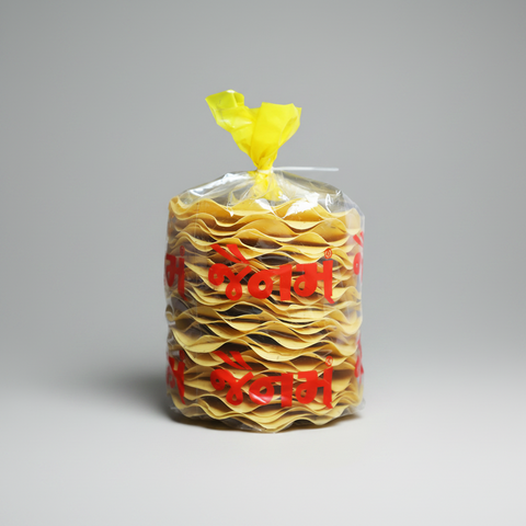 A clear plastic bag filled with stacked yellow flatbread or crackers, tied at the top with a yellow plastic twist, and printed with bold red Devanagari text, shown on a plain white background.
